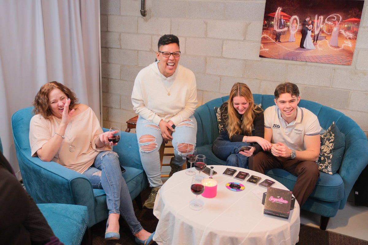 Four people laughing and socializing in a lounge setting, seated on blue armchairs and a sofa around a small round table with drinks and the card game Sexpertease. A framed picture hangs on the brick wall behind them.