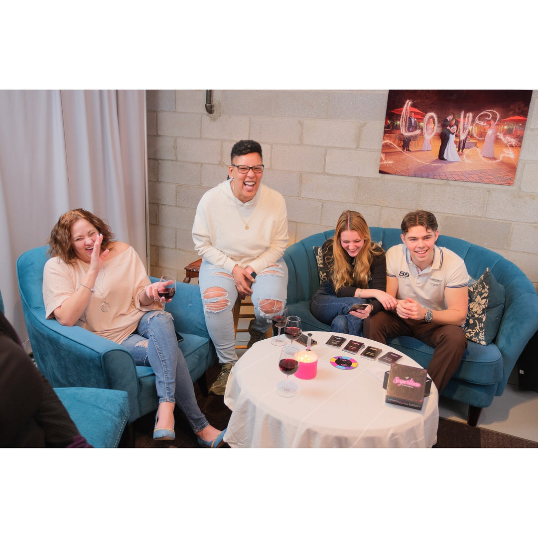 Four people laughing and socializing in a lounge setting with blue armchairs, a round coffee table, and a brick wall background playing the game Sexpertease. They are laughing.